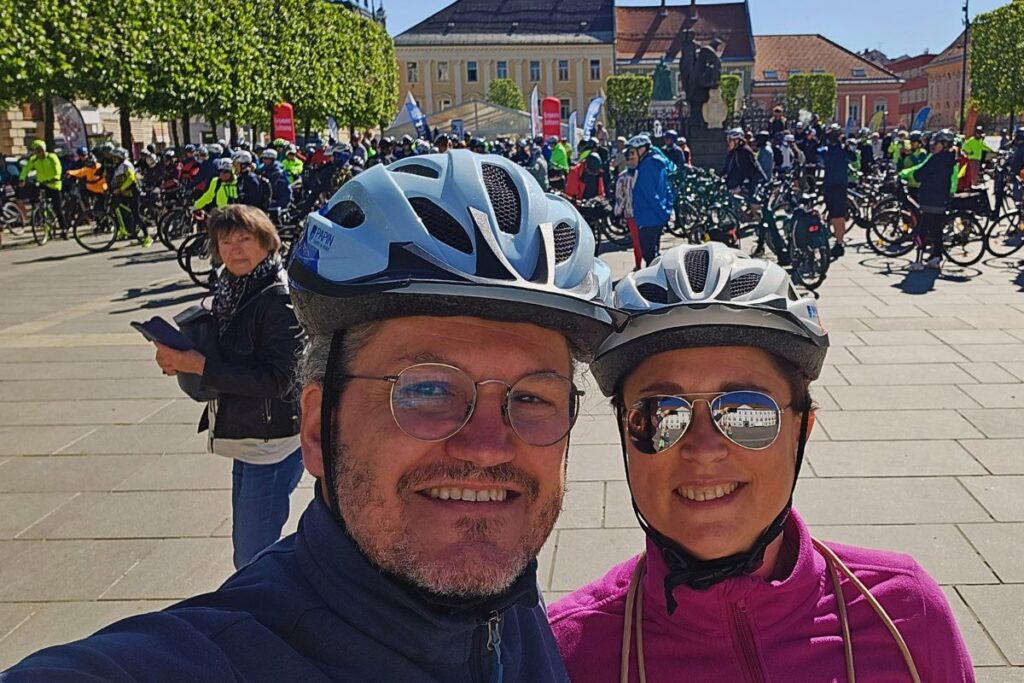 Selfie von Claudia und Nicolo mit Fahrradhelmen und Brillen vor einer großen Menschenmenge mit Fahrrädern beim Etappenstart in Kärnten.