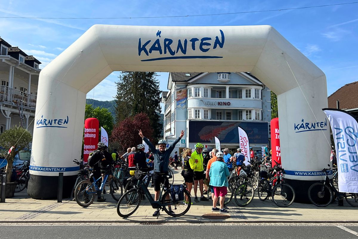Reiseblogger Nicolo Martin jubelt gut gelaunt mit Victory-Zeichen vor einem großen weißen Kärnten-Eventbogen in Pörtschach, im Hintergrund das Wörthersee Kino.