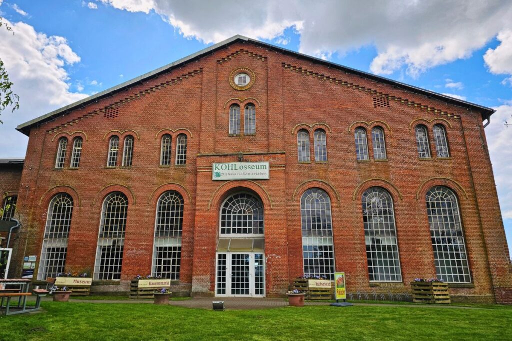 Die imposante historische Backsteinfassade des KOHLosseums in Wesselburen mit großen, hohen Rundbogenfenstern und einem weißen Schild mit der Aufschrift „KOHLosseum – Dithmarschen erleben“ über dem gläsernen Eingang.