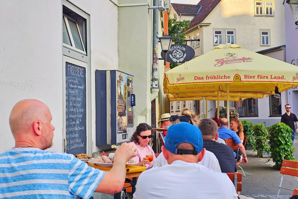 Die Außenterrasse des Biersalons in Coburg mit Gästen an Holztischen unter einem gelben Sonnenschirm und einer handbeschriebenen Tafel mit der Fassbier-Auswahl.