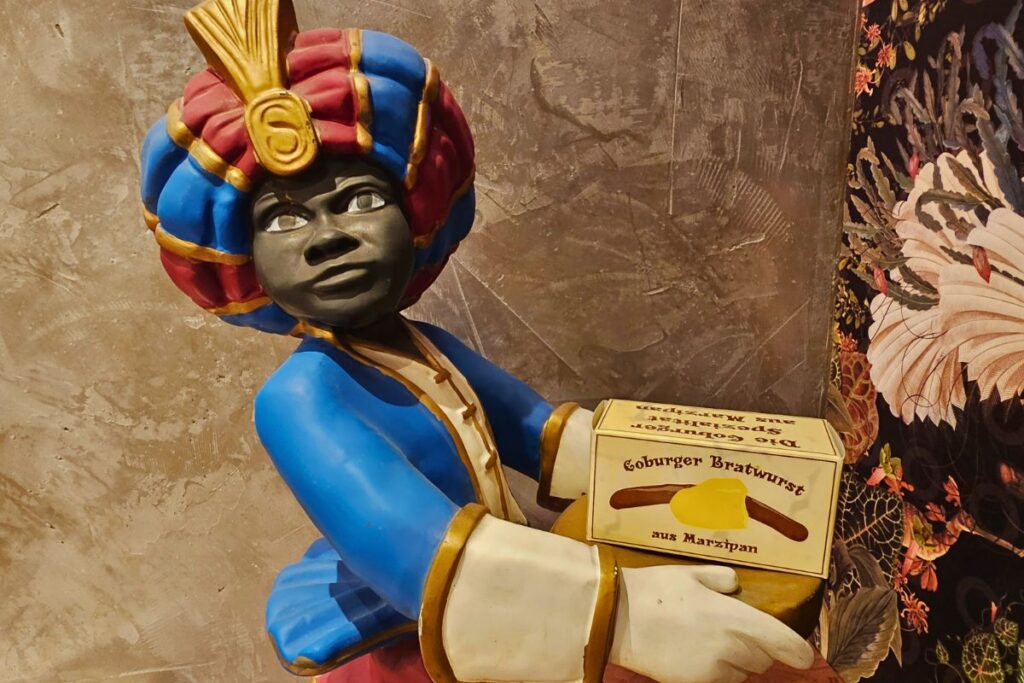 Eine bunte Werbefigur des Coburger Mohrs, die eine Schachtel mit der Aufschrift „Coburger Bratwurst aus Marzipan“ vom Café Schubart präsentiert.
