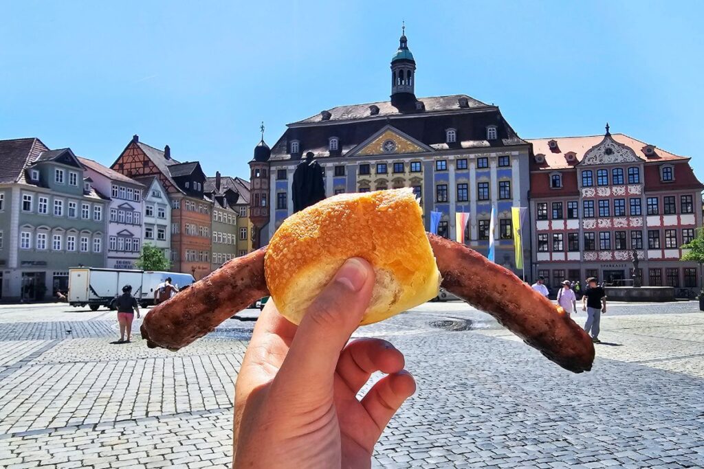 Eine Hand hält eine Coburger Bratwurst in einer kleinen Semmel hoch, im Hintergrund erstreckt sich der kopfsteingepflasterte Marktplatz mit dem historischen Rathaus in Coburg.