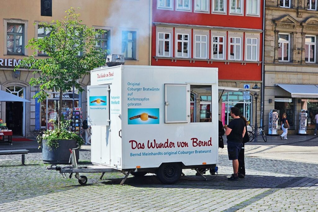 Die weiße mobile Bratwurstbude "Das Wunder von Bernd" auf dem kopfsteingepflasterten Marktplatz in Coburg mit dampfendem Schornstein im Sonnenschein.