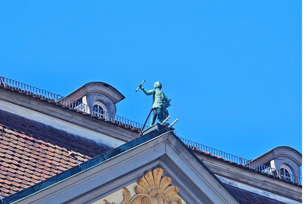 Die historische grüne Kupferstatue des Bratwurstmännla auf dem Dachgiebel des Coburger Rathauses vor strahlend blauem Himmel.