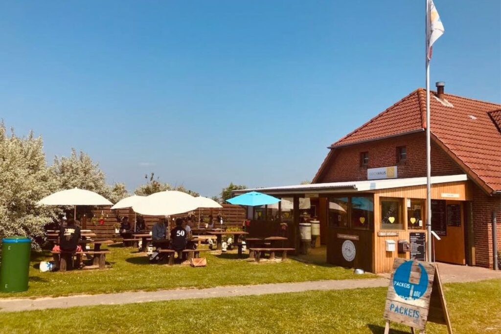 Der sonnige Kaffeegarten des Deichhauses in Dithmarschen mit Holztischen unter weißen Sonnenschirmen vor einem einladenden roten Backsteingebäude.