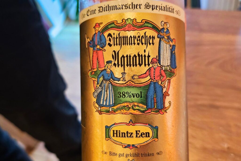 Nahaufnahme des goldenen Etiketts einer Flasche Dithmarscher Aquavit mit der Aufschrift "Hintz Een" und traditionellen norddeutschen Figuren in Tracht.