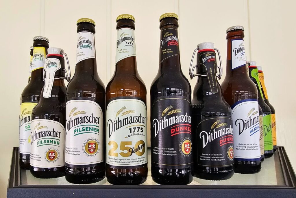 Eine Auswahl verschiedener Bierflaschen der Dithmarscher Privatbrauerei, darunter Pilsener, Dunkel, Radler und alkoholfreies Bier, aufgereiht nebeneinander im Regal.