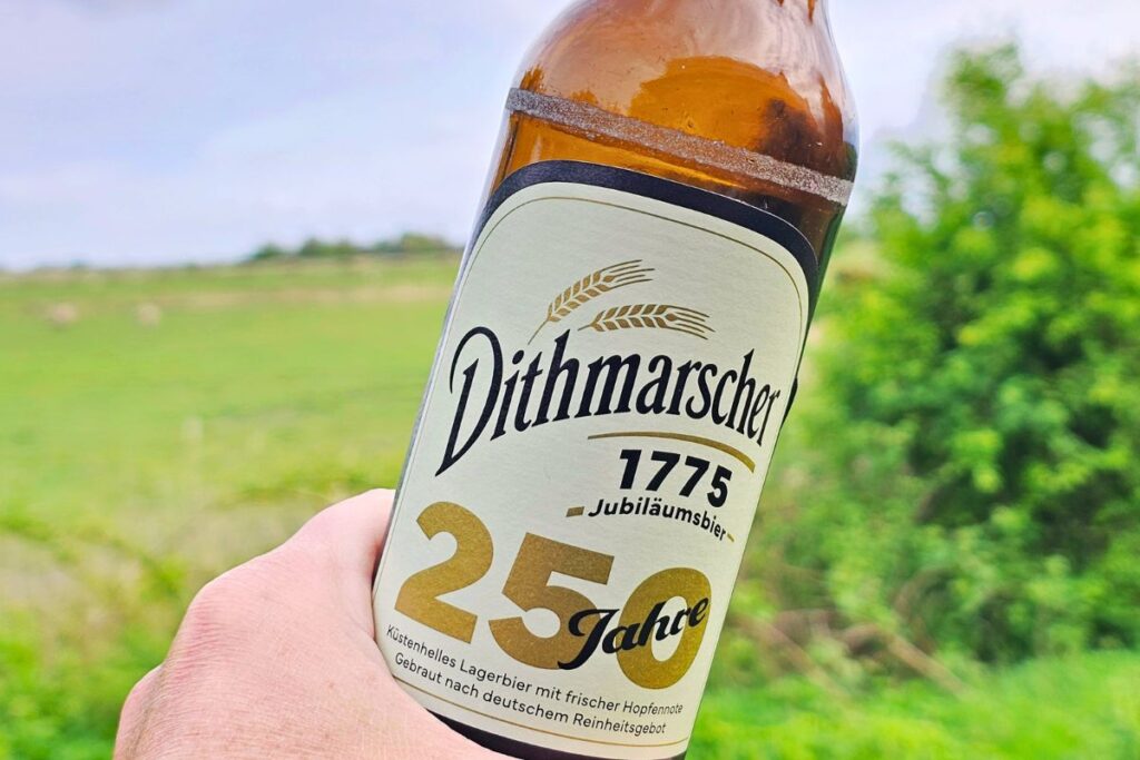 Eine Hand hält eine Flasche Dithmarscher 1775 Jubiläumsbier zum 250-jährigen Bestehen vor einer grünen norddeutschen Wiese unter bewölktem Himmel.