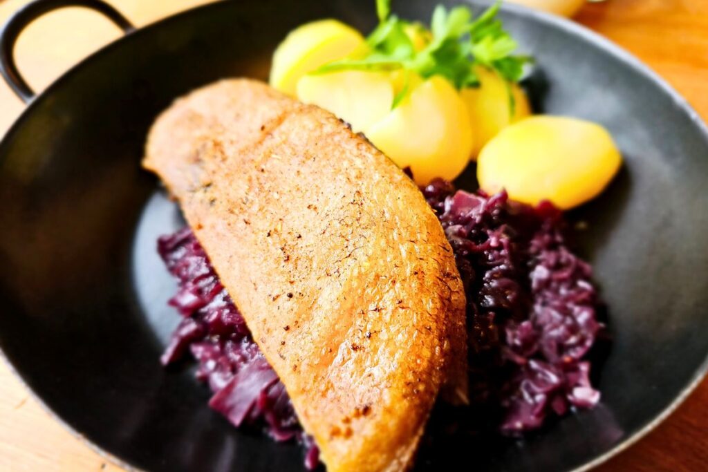 Ein saftiges Stück Gänsebrust serviert auf einer großen Portion Rotkohl mit gekochten Kartoffeln und Petersilie in einer schwarzen gusseisernen Pfanne.