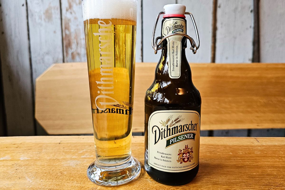 Eine klassische braune Bügelflasche Dithmarscher Pilsener steht neben einem frisch eingeschenkten, schäumenden Bierglas auf einem hellen Holztisch.