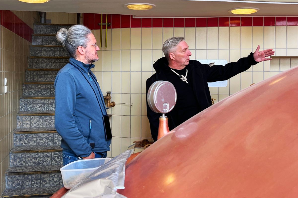 eiseblogger Nicolo Martin steht bei einer Brauereiführung in der Dithmarscher Privatbrauerei in Marne neben Bierlotse Jörg, der auf einen kupfernen Braukessel zeigt.