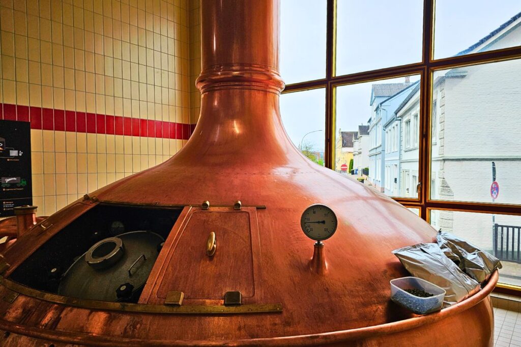 Ein großer, glänzender Braukessel aus Kupfer im Sudhaus der Dithmarscher Privatbrauerei mit Blick durch die großen Fenster auf die Straße von Marne.