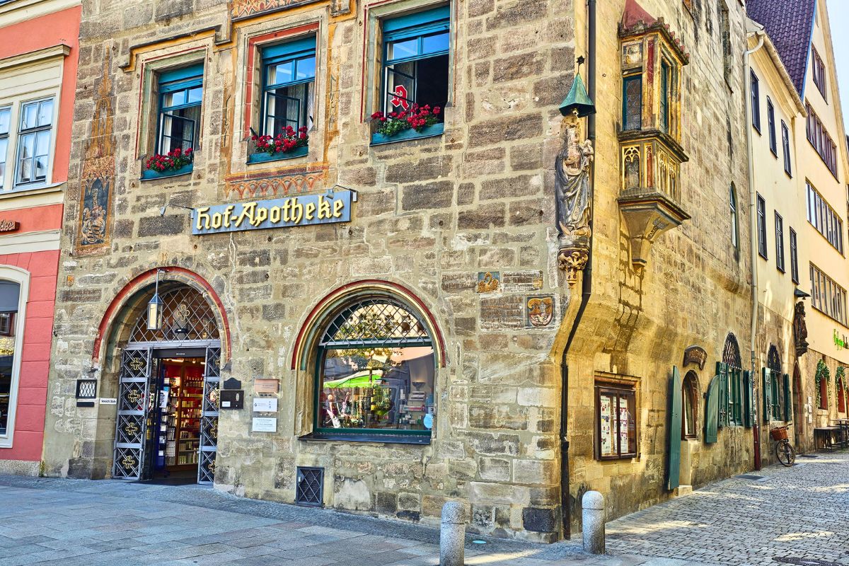 Die beeindruckende historische Steinfassade der Hof-Apotheke in Coburg am Marktplatz mit kunstvollen Malereien, Blumenkästen an den Fenstern und einem Erker.