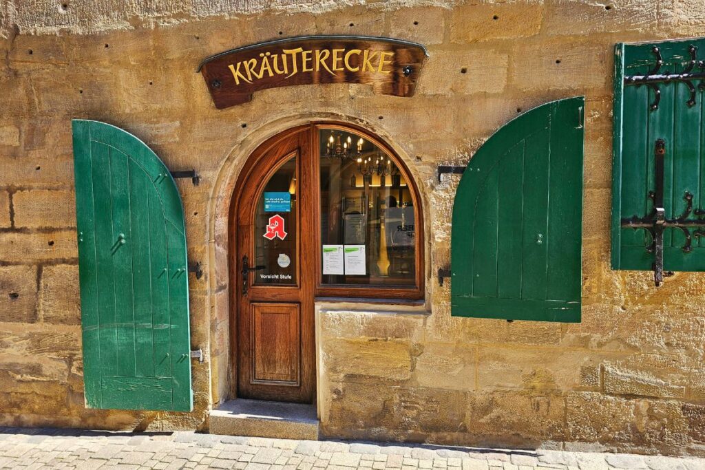 Die historische Sandsteinfassade der Hof-Apotheke am Markt in Coburg mit der Aufschrift „Kräuterecke“ über einer hölzernen Rundbogentür und geöffneten grünen Fensterläden.