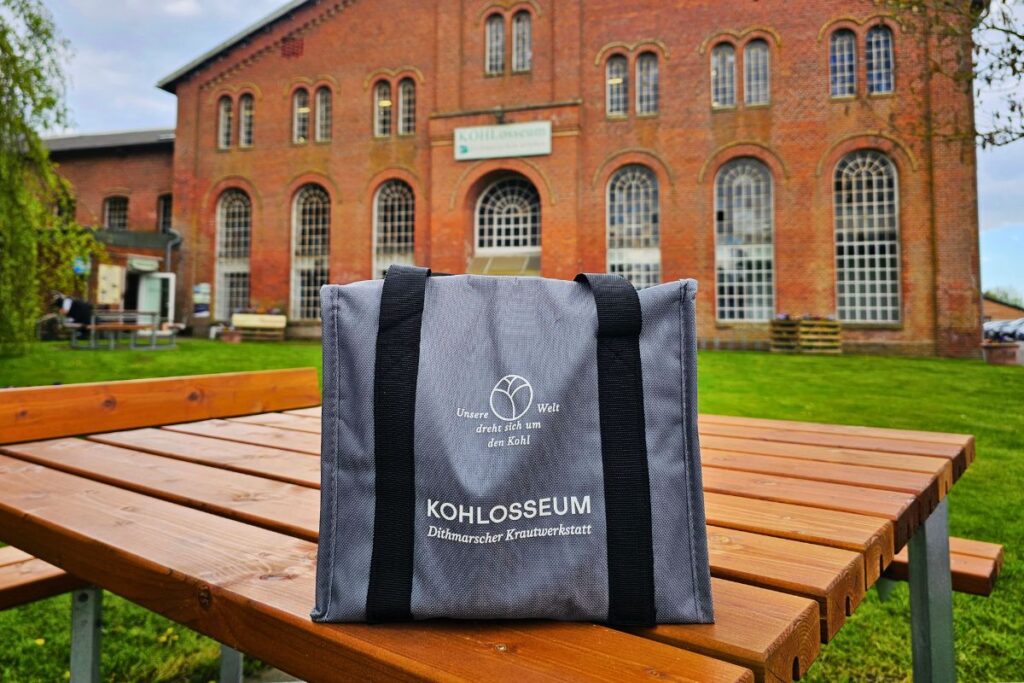 Eine graue Stofftasche mit der Aufschrift „KOHLosseum Dithmarscher Krautwerkstatt“ steht auf einem Holztisch vor dem imposanten historischen Backsteingebäude in Wesselburen.