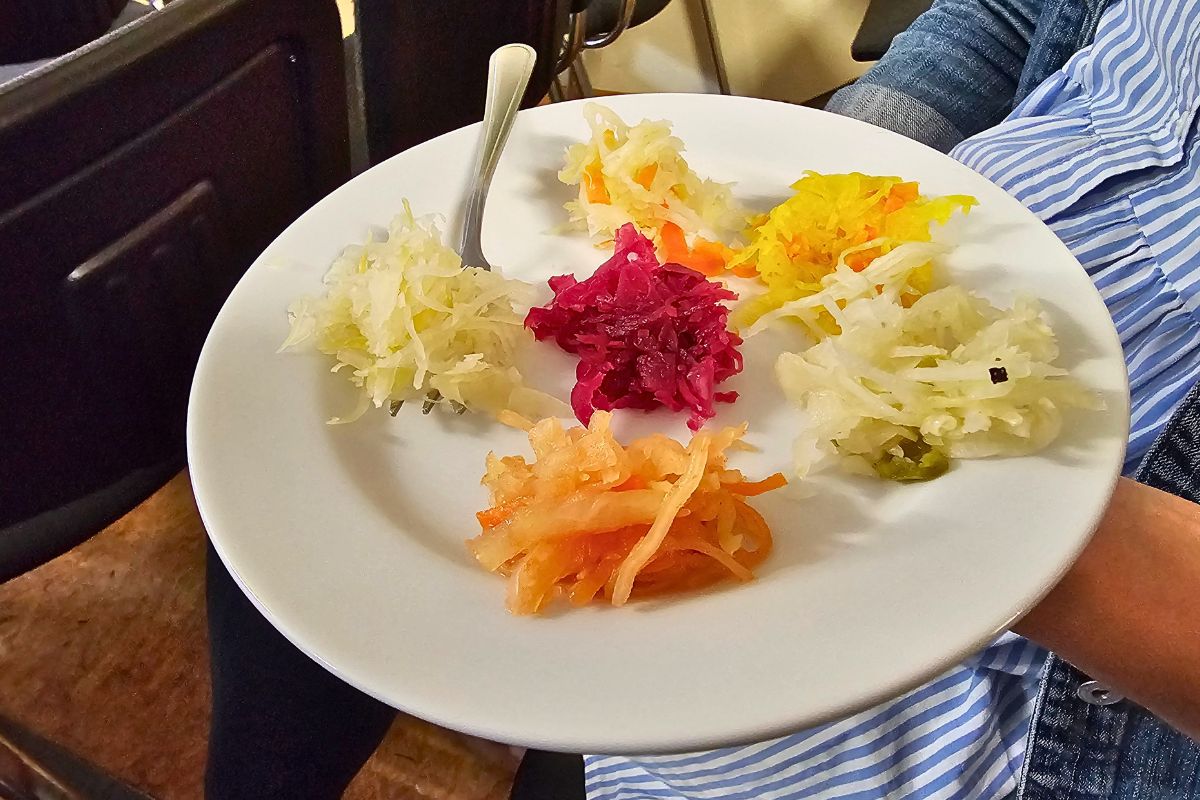 Ein weißer Teller mit sechs verschiedenen, farbenfrohen Sauerkraut- und Krautsalat-Variationen, die zur Verkostung im KOHLosseum Wesselburen gereicht werden.