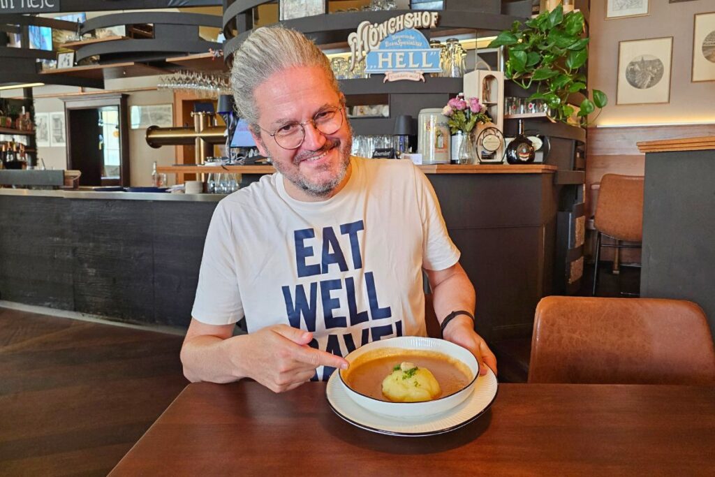 Reiseblogger Nicolo Martin lächelt gut gelaunt im Restaurant in Coburg und zeigt mit dem Finger auf einen weichen Kartoffelkloß (Coburger Rutscher) in reichlich dunkler Bratensoße.