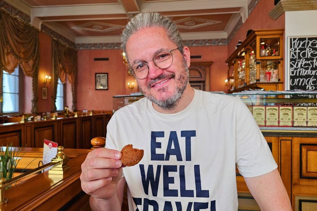 Reiseblogger Nicolo Martin lächelt in die Kamera und hält ein angebissenes Coburger Schmätzchen in der Hand, während er ein weißes T-Shirt mit der Aufschrift „EAT WELL TRAVEL“ trägt.