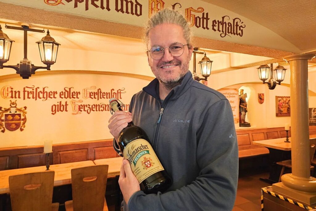 Reiseblogger Nicolo Martin lächelt in die Kamera und hält stolz eine große Flasche Dithmarscher Pilsener im Keller mit Wandinschriften in der Dithmarscher Brauerei.