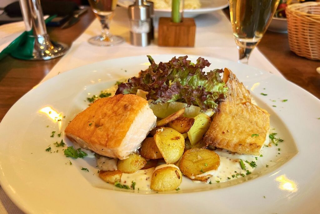 Zwei perfekt kross gebratene Stücke Schollenfilet mit goldbraunen Bratkartoffeln, cremiger Soße und knackigem Beilagensalat auf einem weißen Teller in der Brasserie V Meldorf.