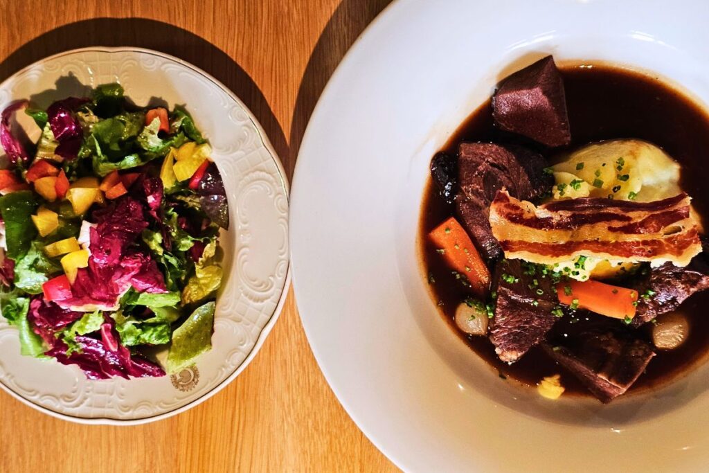 Herzhaftes Dithmarscher Rinderragout in dunkler Soße mit cremigem Kartoffelpüree, Karotten und einer krossen Speckscheibe garniert, daneben ein bunter Beilagensalat im Restaurant Gustav's.