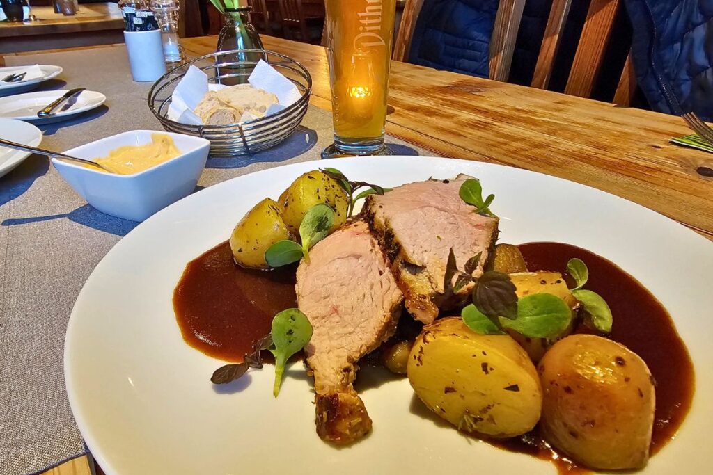 Zarter regionaler Braten in dunkler Soße mit geschwenkten Kräuterkartoffeln und Sprossen auf einem weißen Teller, im Hintergrund ein frisch gezapftes Dithmarscher Pils.