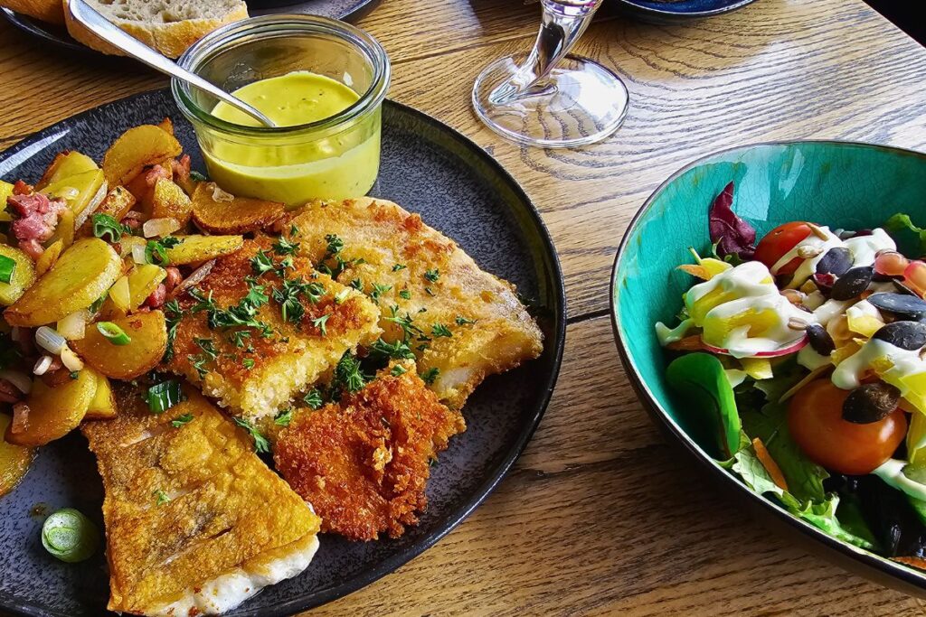 Eine deftige Portion gebratener Fischvariationen mit krossen Bratkartoffeln, Speck und Frühlingszwiebeln neben einer Schale frischem Salat im Restaurant Kohldampfer.
