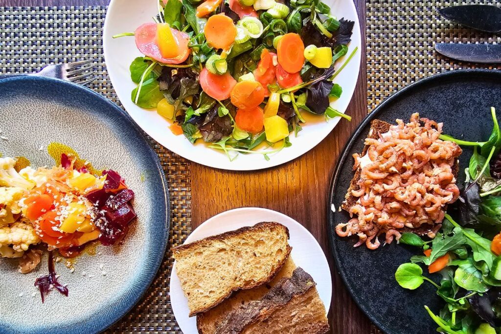 Eine Draufsicht auf reich gedeckte Teller mit frischem Krabbenbrot auf Schwarzbrot, buntem Karottensalat, einer Vorspeise mit Roter Bete und geschnittenem Landbrot.