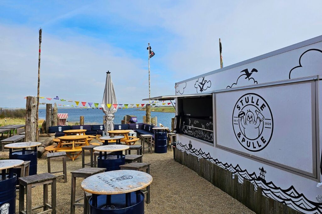 Der weiße Kiosk-Container von Stulle und Pulle mit einer gezeichneten Robbe auf dem Logo, umgeben von Stehtischen aus blauen Ölfässern direkt am Wasser unter blauem Himmel.