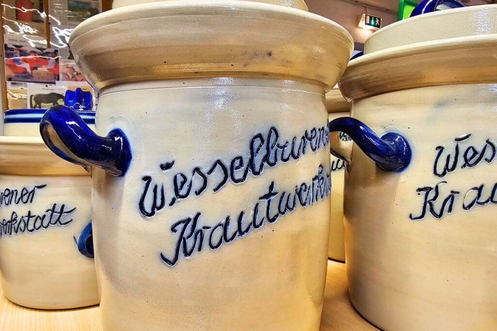 Nahaufnahme von traditionellen beigefarbenen Steinguttöpfen mit blauer Aufschrift „Wesselburen Krautwerkstatt“ und blauen Griffen zur Fermentation von Sauerkraut.