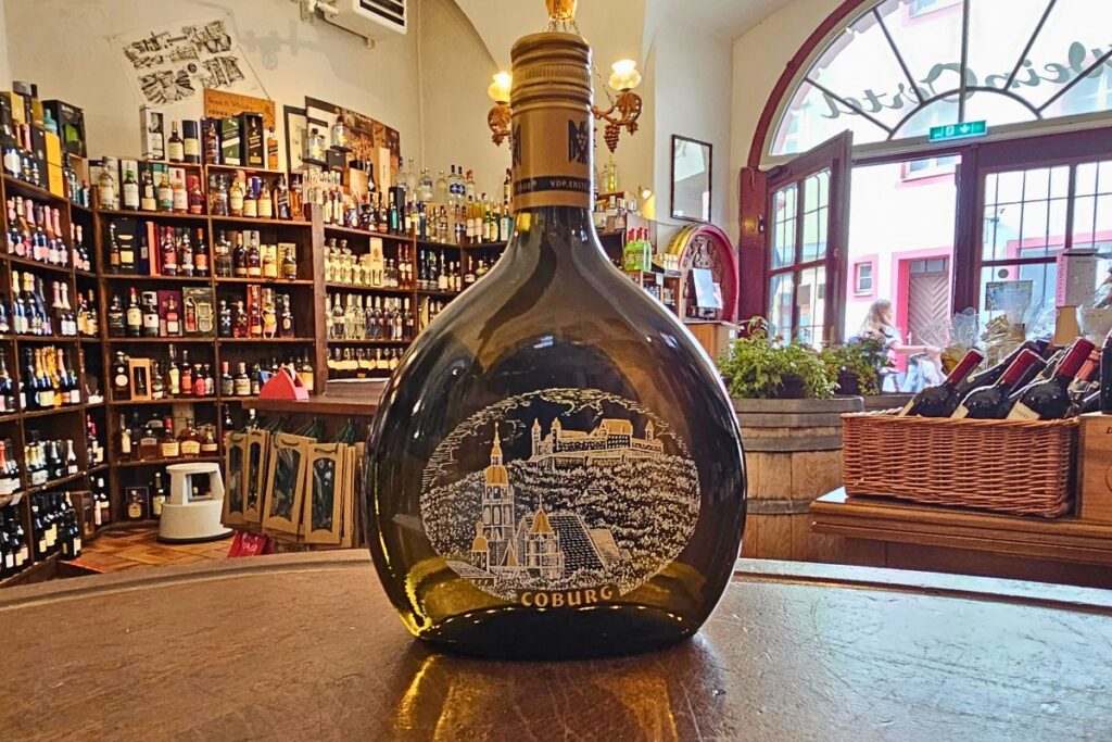 Eine traditionelle, flache Bocksbeutel-Weinflasche von Wein Oertel in Coburg mit einer kunstvollen, weißen Zeichnung der Veste Coburg auf der Vorderseite.