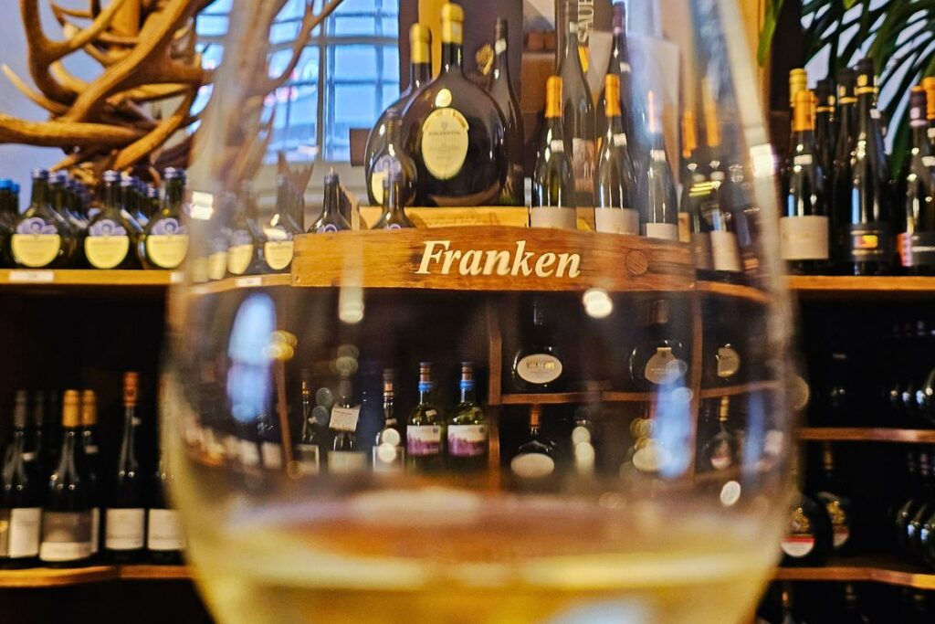 Der stimmungsvolle Blick durch ein Weißweinglas auf die reich gefüllten Holzregale mit dem Schild "Franken" im historischen Verkaufsraum von Wein Oertel.