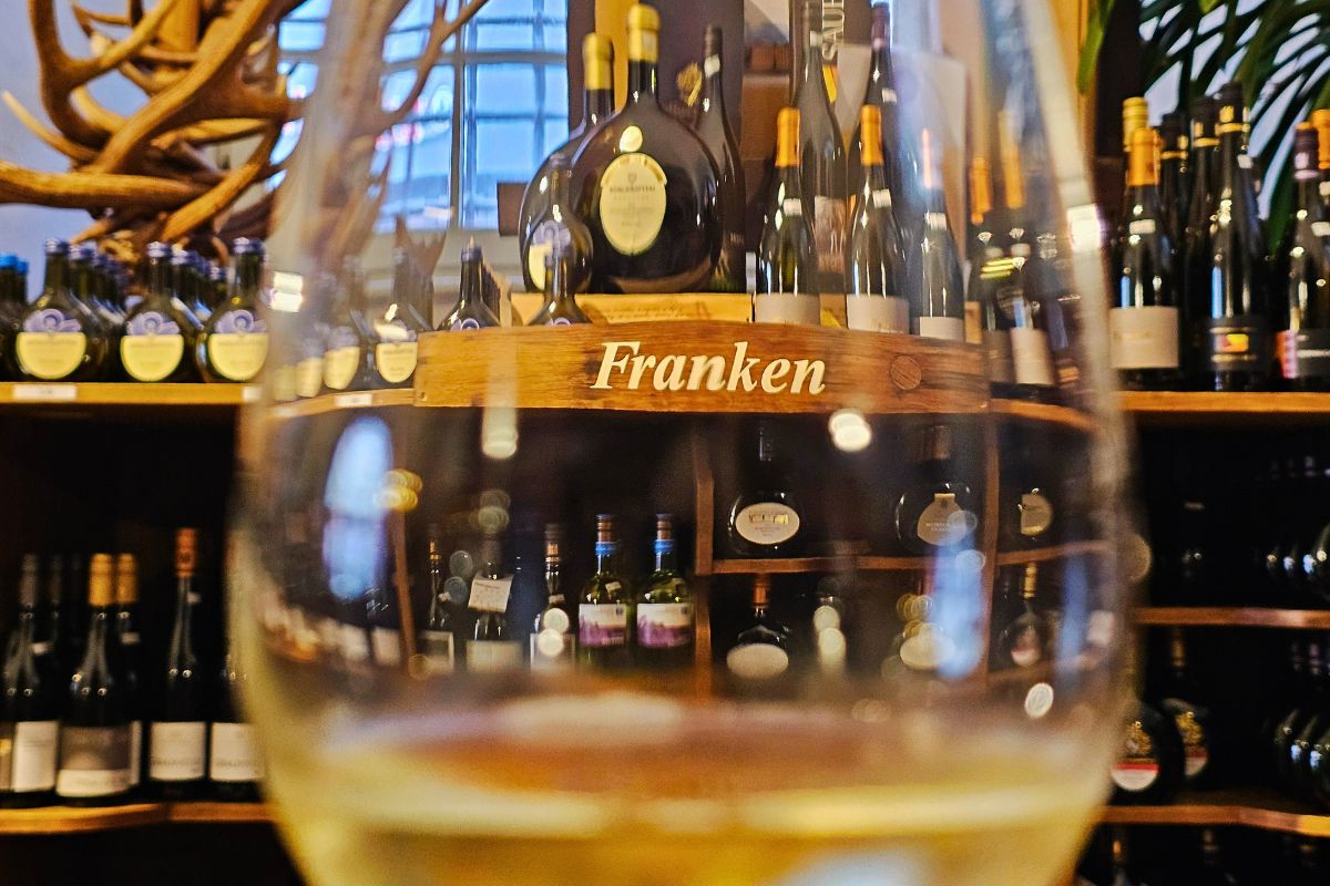 Der stimmungsvolle Blick durch ein Weißweinglas auf die reich gefüllten Holzregale mit dem Schild "Franken" im historischen Verkaufsraum von Wein Oertel.