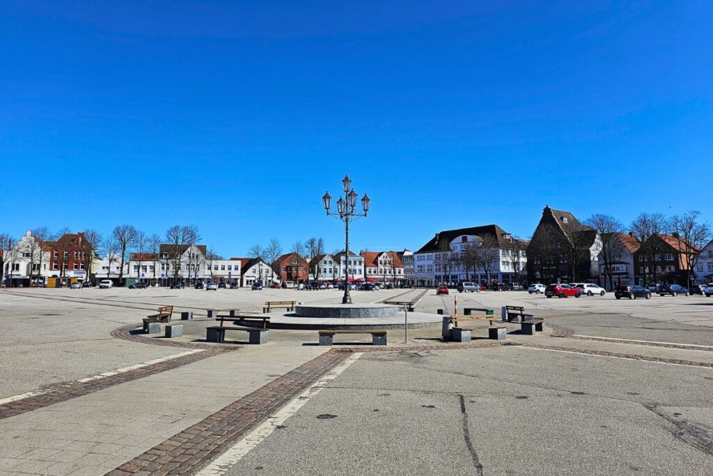Der weitläufige, gepflasterte Marktplatz in Heide mit Bänken und Kandelaber-Laterne im Zentrum unter einem klaren blauen Himmel in Dithmarschen.
