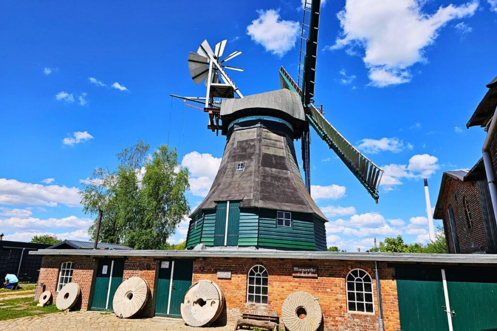 Die historische Galerieholländer-Windmühle Margaretha mit roter Backsteinbasis, grünen Holztoren und alten Mühlsteinen im Vordergrund unter blauem Himmel.