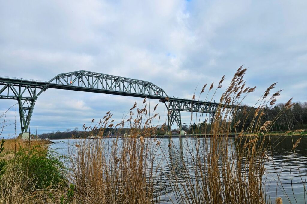 Die monumentale stählerne Eisenbahnhochbrücke Hochdonn überquert den Nord-Ostsee-Kanal, im Vordergrund weht Schilfgras am Ufer unter leicht bewölktem Himmel.