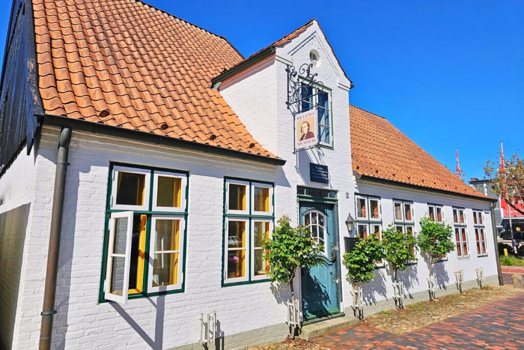 Weiß getünchtes historisches Haus mit rotem Ziegeldach und Auslegerschild des Brahms-Museums auf der Museumsinsel Lüttenheid in Heide.