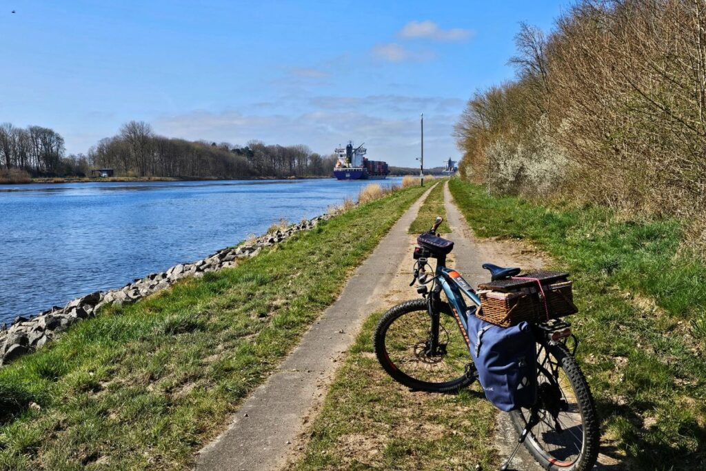 Ein bepacktes Tourenfahrrad steht auf einem asphaltierten Radweg am Ufer des Nord-Ostsee-Kanals, während im Hintergrund ein großes Containerschiff vorbeifährt.