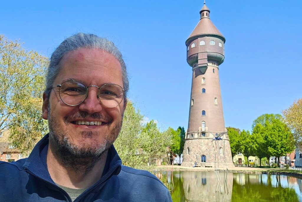 Reiseblogger Nicolo Martin lächelt in die Kamera mit dem historischen, backsteinernen Wasserturm von Heide und einem Teich im Hintergrund.