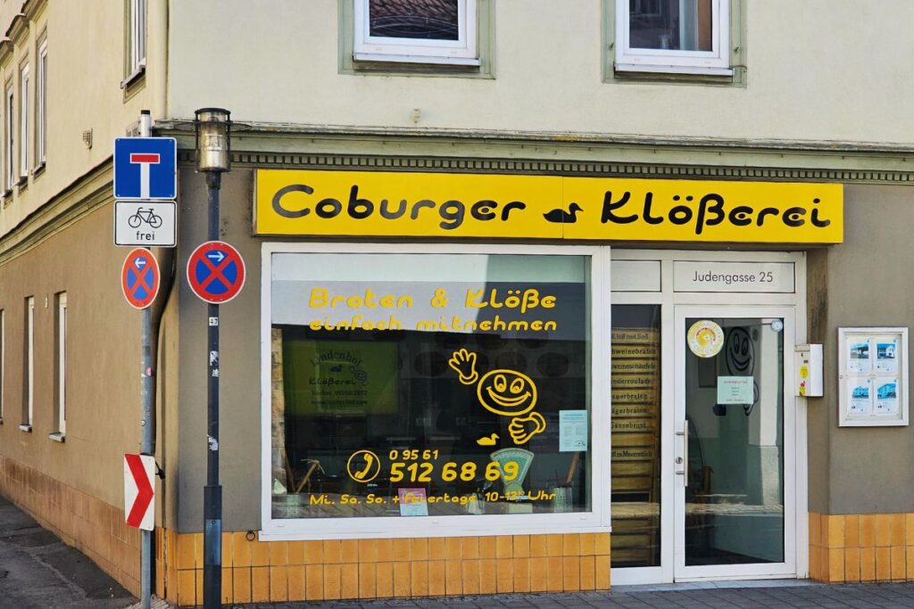 Die Außenfassade der Coburger Klößerei in der Judengasse 25 mit gelbem Firmenschild und Schaufensteraufschrift für Klöße und Braten zum Mitnehmen.