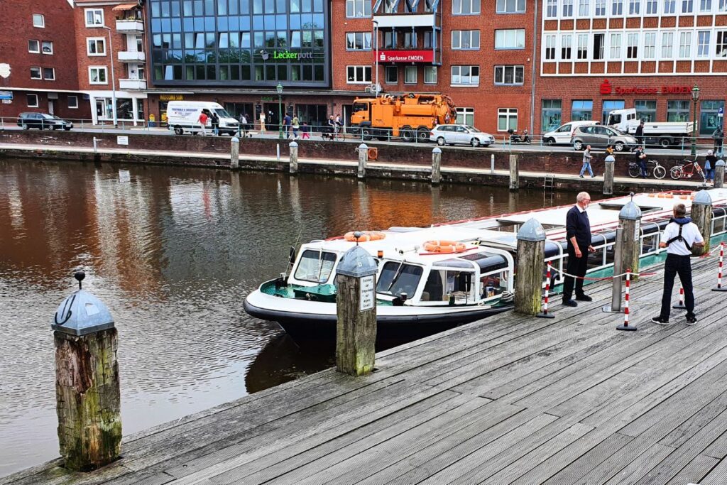 Ein flaches Ausflugsboot für Grachtenfahrten liegt an einem Holzsteg im Emder Ratsdelft vor den Backsteingebäuden der Innenstadt.