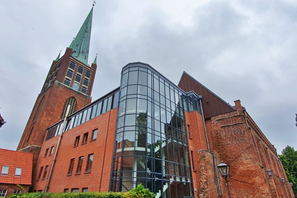 Historischer Kirchturm und rote Backsteinruine der Johannes a Lasco Bibliothek in Emden mit modernem Glasanbau vor wolkigem Himmel.