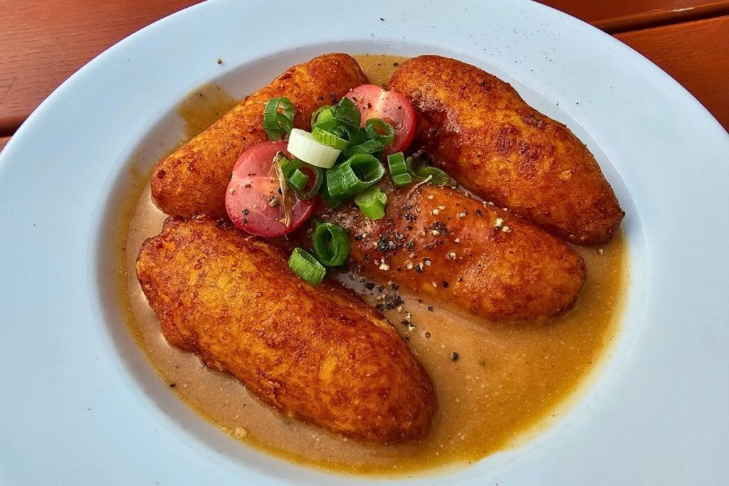 Vier kross gebratene, längliche Kloßstäbla in sämiger Sauce, garniert mit Frühlingszwiebeln, Minitomaten und grobem Pfeffer im Biersalon Coburg.
