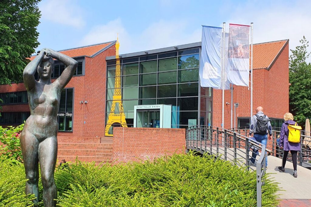 Der Backsteinbau der Kunsthalle Emden mit einer bronzenen Frauenstatue im Vordergrund, Glasfront und Besucherbrücke.