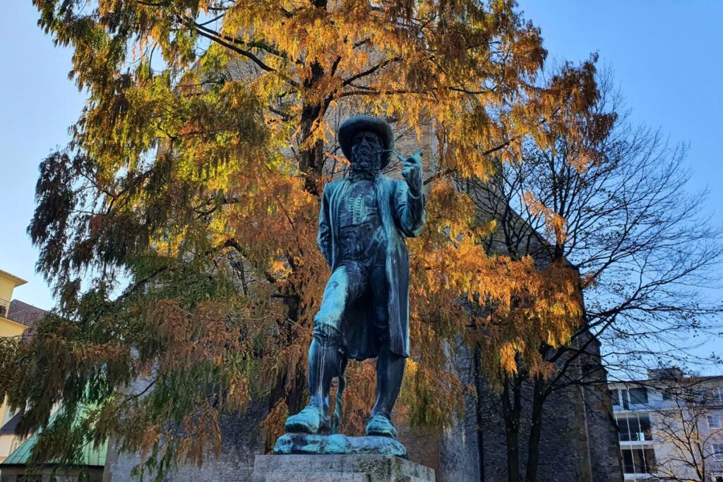 Das bronzene Leineweberdenkmal mit Tabakspfeife und Leinenballen vor einem herbstlich gefärbten Baum auf dem Altstädter Kirchplatz in Bielefeld.