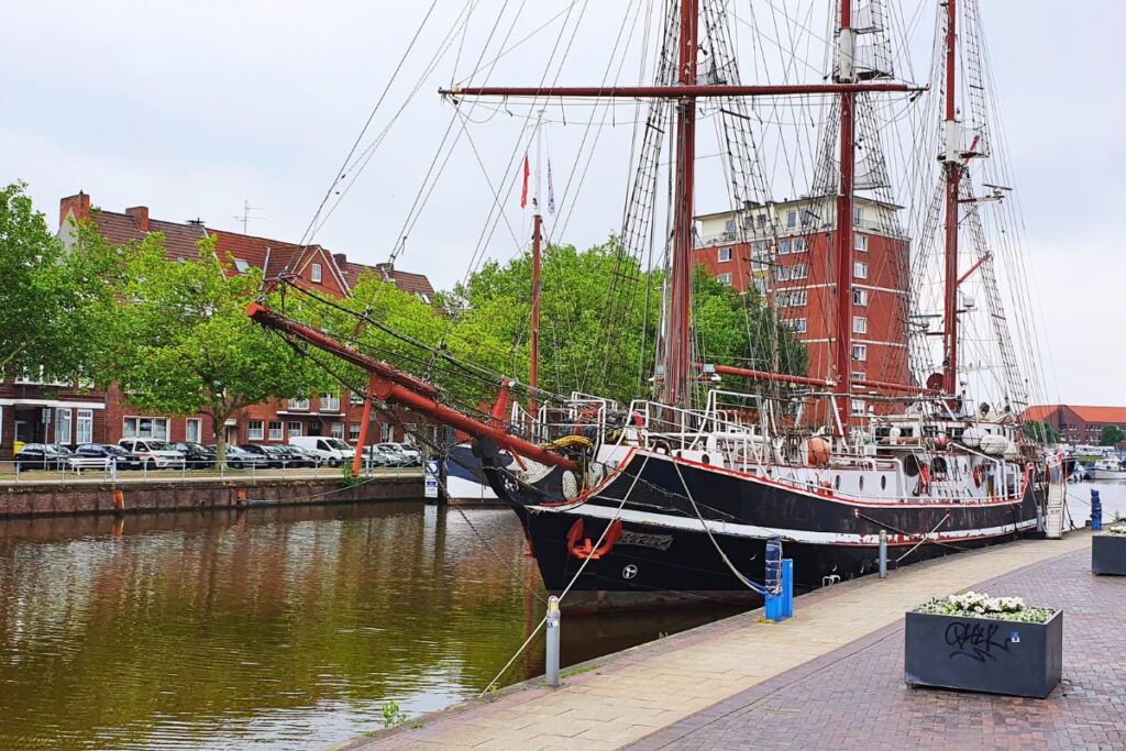 Ein historischer Traditionssegler mit dunklem Holzrumpf und Takelage liegt vertäut im Ratsdelft vor Backsteinhäusern in Emden.