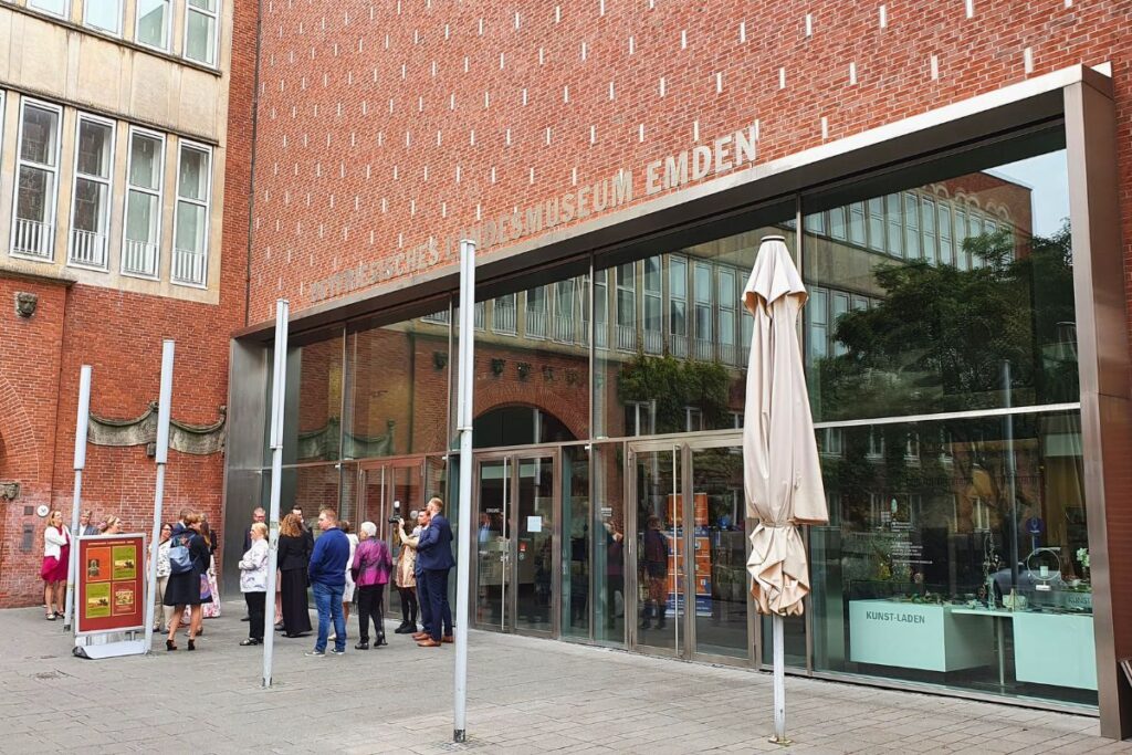 Die moderne Glasfront und Backsteinfassade des Ostfriesischen Landesmuseums im Rathaus Emden mit einer Gruppe wartender Besucher auf dem Vorplatz.