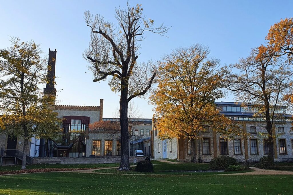 Das restaurierte Backsteingebäude der Ravensberger Spinnerei mit Schornstein und herbstlicher Parkanlage in Bielefeld.