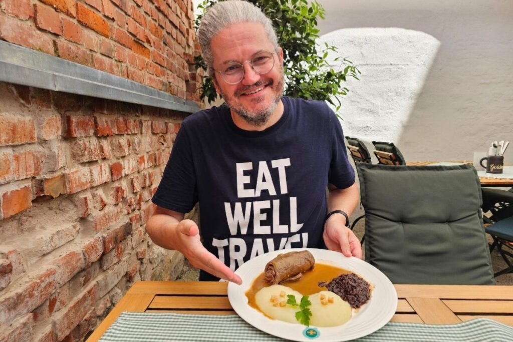 Reiseblogger Nicolo Martin lächelt im Innenhof des Restaurants Goldenes Kreuz und präsentiert einen Teller mit Rinderroulade, zwei Coburger Rutschern und Rotkohl.