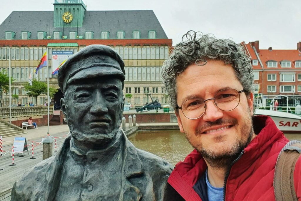 Reiseblogger Nicolo Martin lächelt neben einer der bronzenen Seemannsskulpturen der Delftspucker am Ratsdelft vor dem historischen Emder Rathaus.